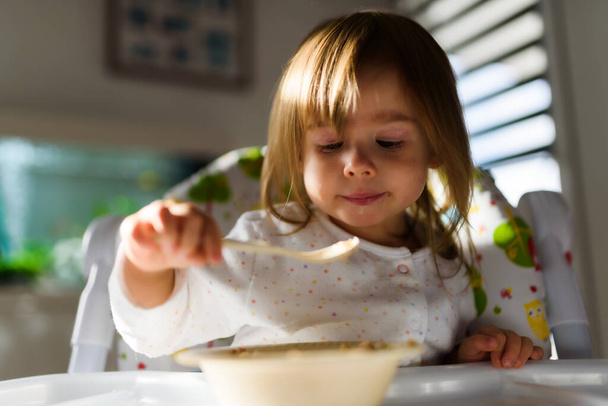 Two years old eats brakefast by herself with a spoon. Child development concept - Φωτογραφία, εικόνα