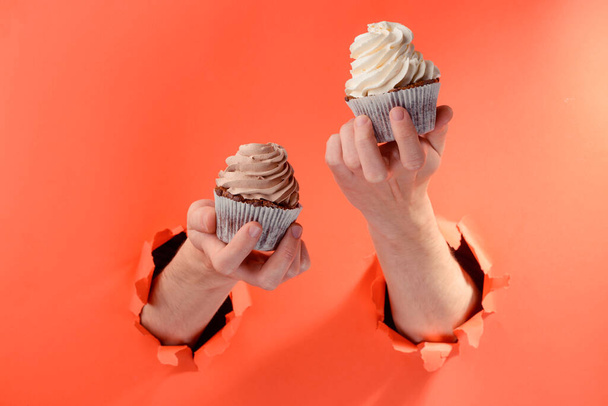 Hands holding two cupcakes through torn holes in red paper background - Φωτογραφία, εικόνα