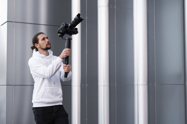 Νέοι επαγγελματίες videographer κρατώντας επαγγελματική φωτογραφική μηχανή σε 3-άξονα gimbal σταθεροποιητή. Pro εξοπλισμός βοηθά να κάνει υψηλής ποιότητας βίντεο χωρίς ανακίνηση. Καμεραμάν με άσπρη κουκούλα που φτιάχνει βίντεο. - Φωτογραφία, εικόνα