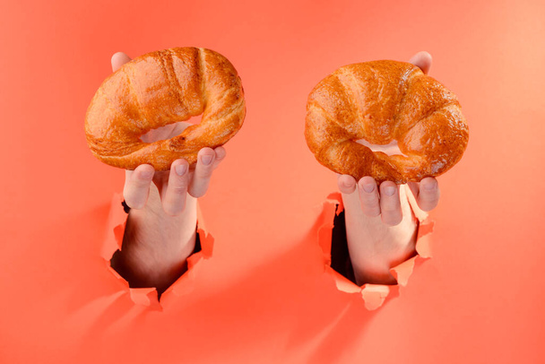 Hands holding two croissants through torn holes in red paper background - Fotografie, Obrázek