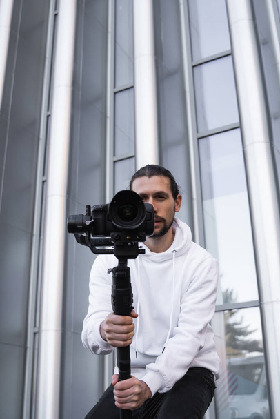 Jonge professionele videograaf met professionele camera op 3-assige gimbal stabilisator. Pro-apparatuur helpt om video van hoge kwaliteit te maken zonder te schudden. Cameraman draagt witte hoodie een video 's maken. - Foto, afbeelding