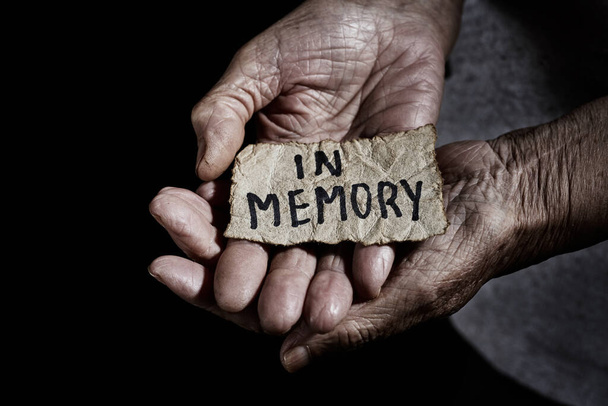 closeup of a yellowish piece of paper, with the text in memory written in it, in the wrinkled hands of a senior woman, with a dramatic light - Photo, Image