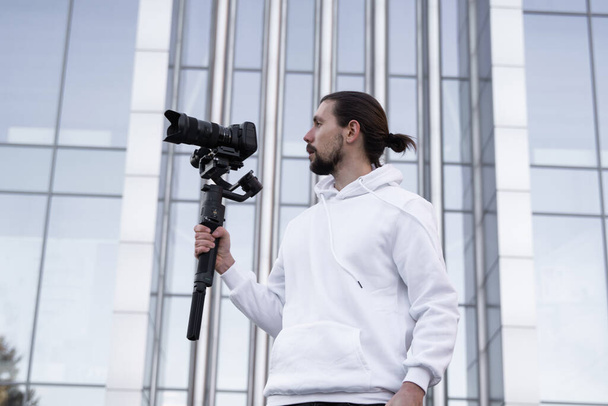 Young Professional videographer holding professional camera on 3-axis gimbal stabilizer. Pro equipment helps to make high quality video without shaking. Cameraman wearing white hoodie making a videos. - Zdjęcie, obraz