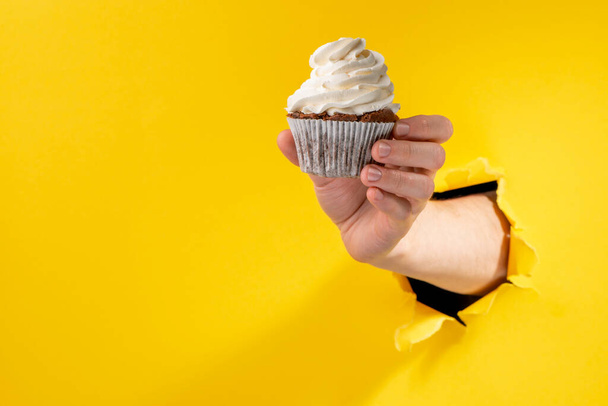 Hand giving a cupcake through torn yellow paper background - Fotó, kép