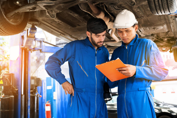 De monteur in blauw werkuniform inspecteert de onderkant van de auto met zijn assistent. Automobiel reparatie service, Professioneel beroep teamwork. Onderhoud van voertuigen. - Foto, afbeelding
