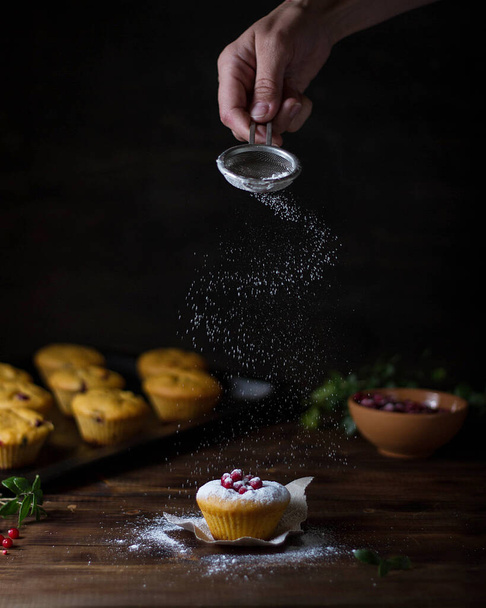 Pumpkin muffins with cranberries on a dark wooden table. A hand with a strainer pours powdered sugar on top. - 写真・画像