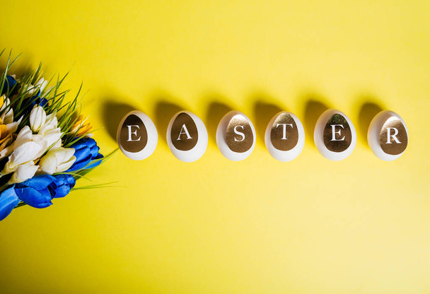 Easter concept. White eggs, golden letters and a bouquet of flowers on a yellow background. Top view - Foto, afbeelding