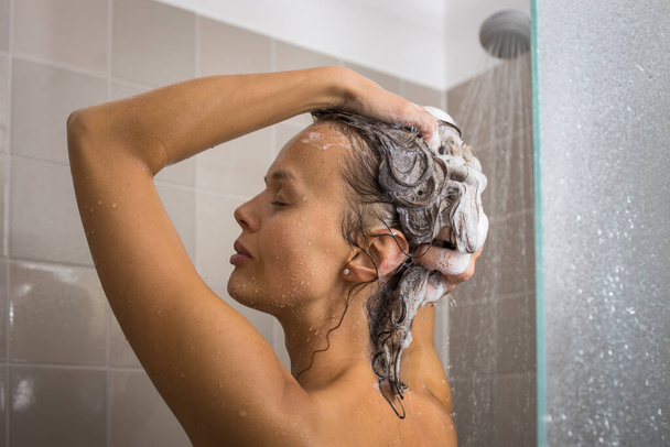 mooie, jonge vrouw het nemen van een lange warme douche wassen haar haar in een modern design badkamer - Foto, afbeelding