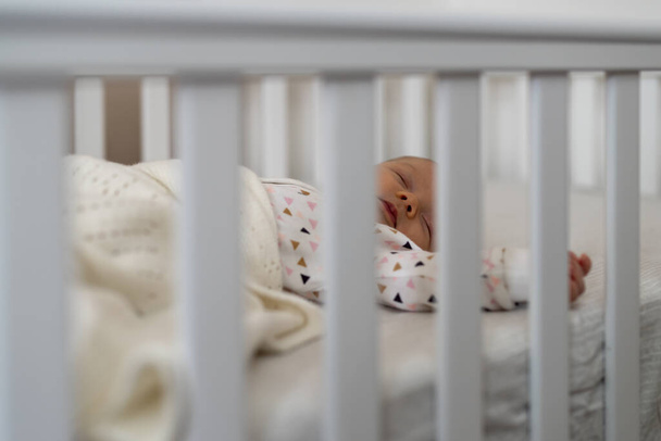 素敵な新生児の女の子はベッドで寝て、ベビーベッドバーを通して見る - 写真・画像