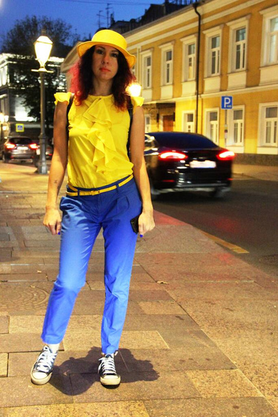 Μια βόλτα στη νυχτερινή πόλη. Μια γυναίκα με κόκκινα σγουρά μαλλιά, κίτρινο καπέλο, μπλε παντελόνι και αθλητικά παπούτσια. κίτρινο-μπλε φανταχτερό κοστούμι, βραδινό φως. τόνωση - Φωτογραφία, εικόνα