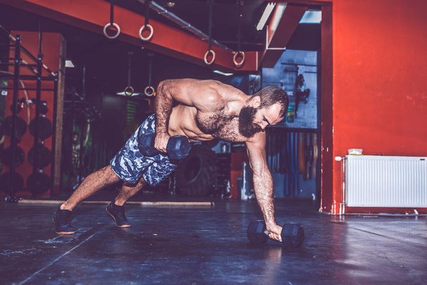 Muscular bearded man with naked torso doing push up exercise with dumbbell in the gym. - Foto, Imagem