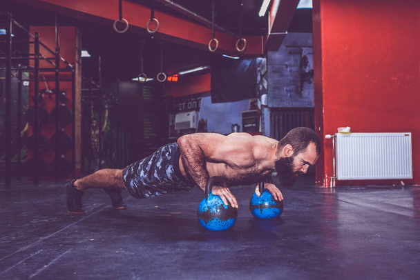 Junger bärtiger Mann mit nacktem Oberkörper macht Liegestütze mit Kettlebell-Gewichten im Fitnessstudio - Foto, Bild