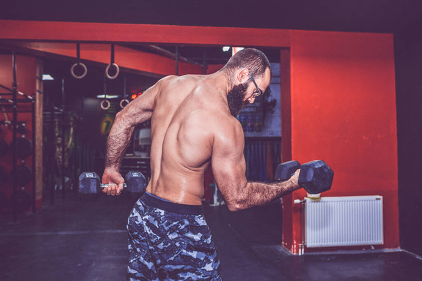 Young bearded man, with naked torso doing exercise and lifting dumbbell weights in gym - Photo, Image