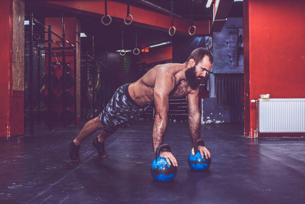 Joven hombre barbudo muscular con el torso desnudo haciendo flexiones con pesas kettlebell en el gimnasio
 - Foto, Imagen