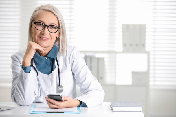 Mature female doctor with smartphone at table in office - Valokuva, kuva