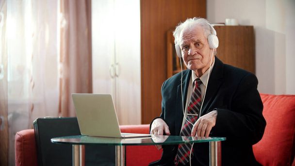An old man in headphones sitting by the laptop - Фото, изображение