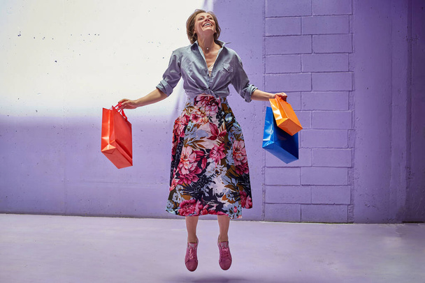 Young beautiful girl jumping of joy with shopping in her hands. A girl in a long bright skirt and pale pink top is walking from the store. - Foto, imagen