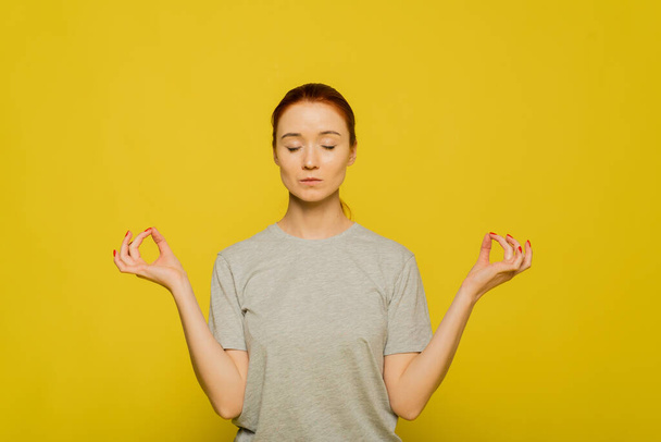 Yoga and meditation. Beautiful casually dressed young redhead woman keeping eyes closed while meditating, feeling relaxed, calm and peaceful after hard working day at office, holding hands in mudra sign. - Valokuva, kuva