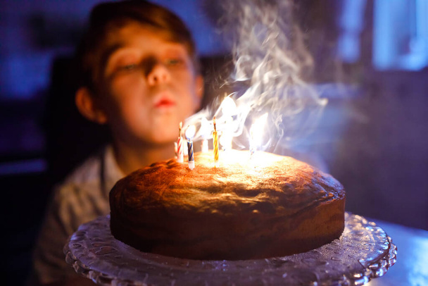Schattige blonde kleine jongen die zijn verjaardag viert. Kind blaast kaarsen op zelfgebakken taart, binnen. Verjaardagsfeest voor schoolkinderen, familiefeest - Foto, afbeelding