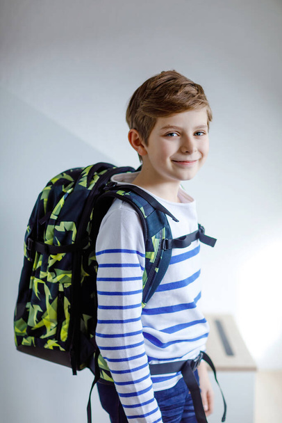 Beautiful kid boy with schoolbag rucksack standing in room in early morning. Happy healthy child on the way to lessons and school. Education concept - Zdjęcie, obraz