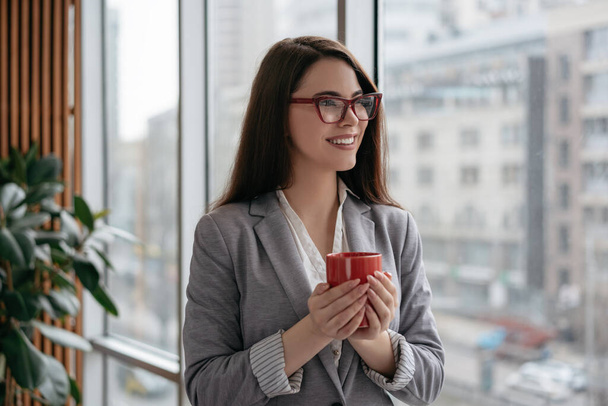 Portret szczęśliwej kobiety w formalnych ubraniach i stylowych okularach planujących start. Młoda, pewna siebie bizneswoman stojąca w nowoczesnym biurze przy oknie, trzymająca filiżankę kawy i uśmiechnięta - Zdjęcie, obraz