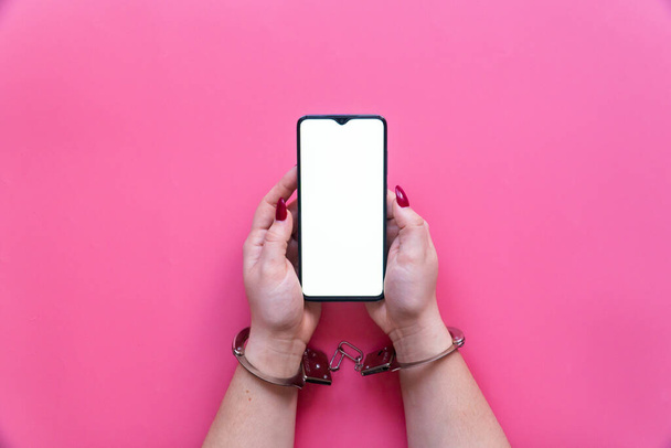 Woman hands in handcuffs hold a smartphone with blank screen on a pink background. The concept of internet and gadget dependency. - Φωτογραφία, εικόνα
