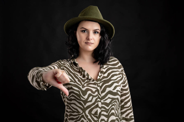 Young woman dressed casually in an animal print shirt, with black hair wearing a hat showing dislike posing on a black isolated backround. - Photo, Image