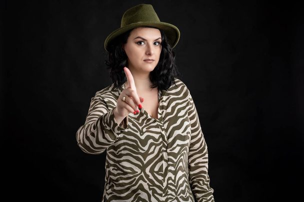Mujer joven vestida casualmente con una camisa de estampado animal, con el pelo negro usando un sombrero que muestra no con el dedo posando sobre un fondo negro aislado
. - Foto, Imagen
