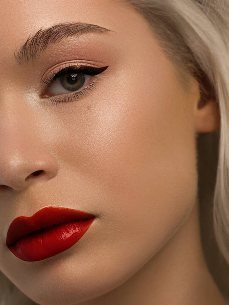 Close-up beauty of half female face with creative fashion evening make-up. Black arrows on the eyes and extremely long eyelashes, on plump lips matte scarlet lip color. Well-groomed skin after spa - Photo, Image