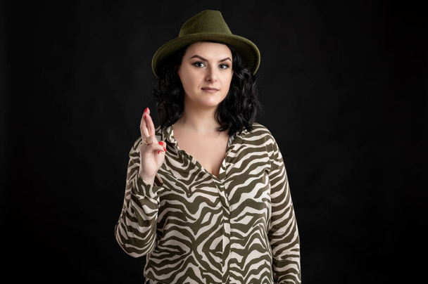 Mujer joven vestida casualmente con una camisa de estampado animal, con el pelo negro usando un sombrero que muestra buena suerte posando sobre un fondo negro aislado
. - Foto, Imagen