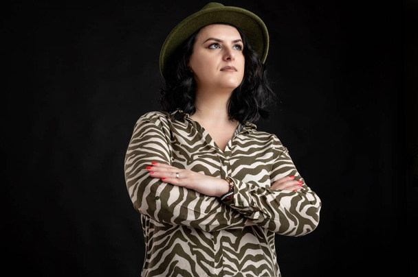 Jonge vrouw gekleed terloops in een dier print shirt, met zwart haar dragen van een hoed ziet er zelfverzekerd held-shot poseren op een zwarte geïsoleerde back-round. - Foto, afbeelding