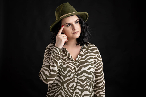 Young woman dressed casually in an animal print shirt, with black hair wearing a hat, front view portrait of a woman wondering, posing on a black isolated backround. - Photo, Image