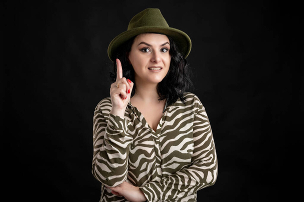Mulher jovem vestida casualmente em uma camisa de impressão animal, com o cabelo preto usando um chapéu com o dedo para cima surgiu com uma ideia posando em um fundo isolado preto
. - Foto, Imagem