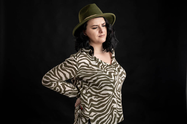 Young woman dressed casually in an animal print shirt, with black hair wearing a hat has back pain posing on a black isolated backrAttractive young woman dressed casually has back painound. - Photo, Image