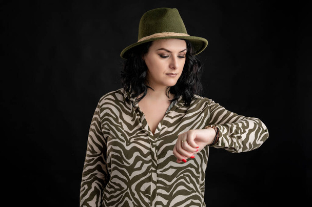 Mujer joven vestida casualmente con una camisa de animal print, con el pelo negro con un sombrero mirando su reloj, es hora de posar sobre un fondo negro aislado
. - Foto, imagen
