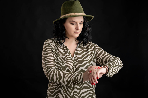 Jonge vrouw gekleed terloops in een dier print shirt, met zwart haar dragen van een hoed kijken haar horloge, het is tijd poseren op een zwarte geïsoleerde back-round. - Foto, afbeelding