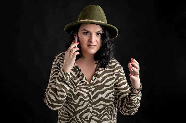 Mujer joven vestida casualmente con una camisa de estampado animal, con el pelo negro con un sombrero disgustado cara y gestos y hablando en el teléfono celular posando en un fondo aislado negro
. - Foto, imagen