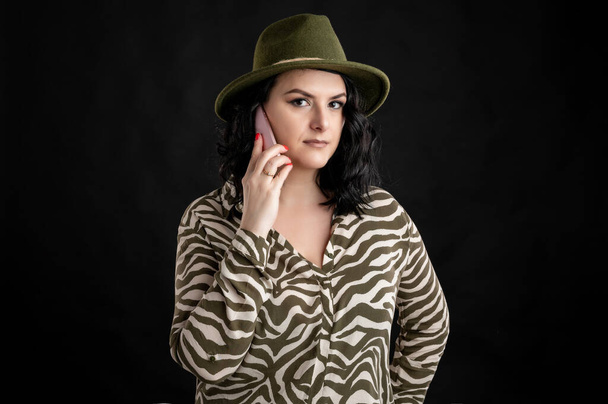 動物のプリントシャツに身を包んだ若い女性、携帯電話で帽子をかぶる黒い髪、黒い隔離された背景にポーズをとるスマートフォン. - 写真・画像