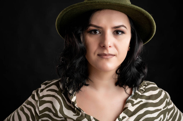 Young woman dressed casually in an animal print shirt, with black hair wearing a hat close-up portrait posing on a black isolated backround. - Photo, Image