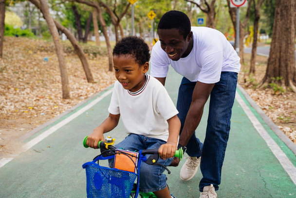 Smiling Happy African American father helps his son learn cycling on bicycle in park. - Foto, Imagen