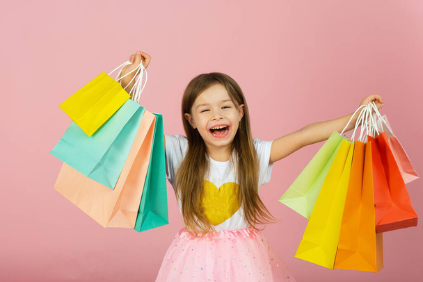 Little girl with many shopping bags on pastel pink backgound. Pretty joyful young girl in tulle skirt, with long blonde hair walking with colorful packages on pink background. - Foto, afbeelding