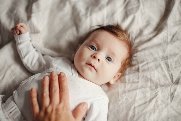 Closeup portrait of cute Caucasian newborn baby. Adorable funny child infant with blue grey eyes and red hair lying on bed looking at camera. Authentic childhood and lifestyle candid moment. - 写真・画像