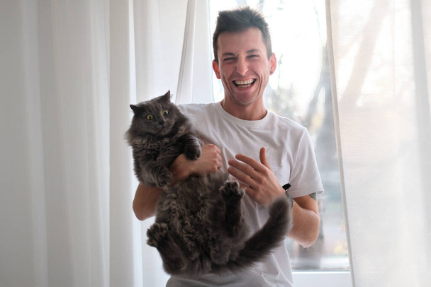 Young man in the white shirt and pajama is smiling holding fluffy cat which wants to run away - Fotoğraf, Görsel