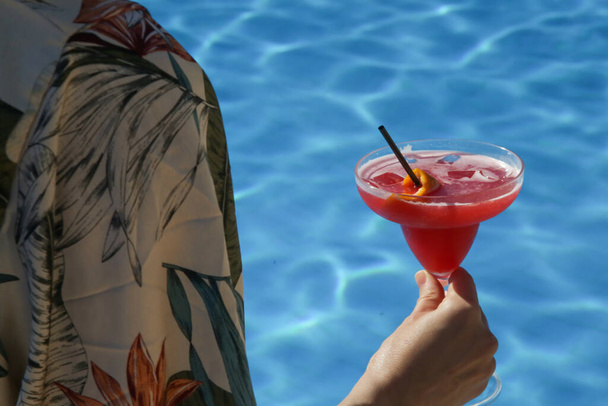 Havuz kenarında oturan ve elinde kokteyl bardağı tutan genç bir kadın, yaz tatili eğlence aktivitesi. - Fotoğraf, Görsel