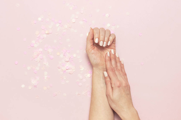 Stylish fashionable female manicure in pastel colors. Hands of young girl on pink background with festive confetti. Minimalist manicure trend. Flat lay, top view, copy space. Natural nails, gel polish. - Photo, image