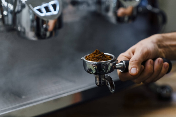 Λεπτομέρεια του ανθρώπου χέρι κρατώντας portafilter παραλήπτης γεμάτο με κόκκους καφέ κατά την προετοιμασία espresso - Φωτογραφία, εικόνα