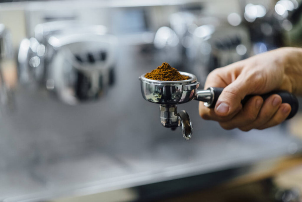 Detalle de la mano del hombre sosteniendo portafilter recipiente lleno de café molido mientras se prepara espresso
 - Foto, imagen