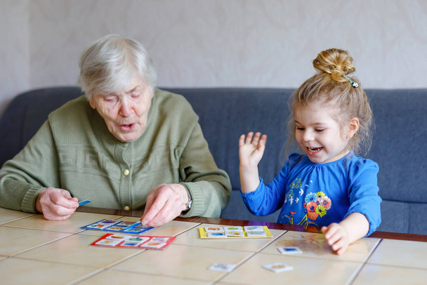 Krásné batole dívka a prababička hrát společně obrázky lotto stolní karty doma. Roztomilé dítě a starší žena se spolu baví. Šťastná rodina uvnitř - Fotografie, Obrázek