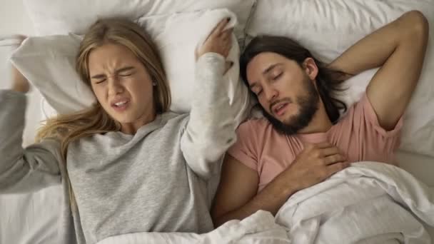 A displeased disappointed young woman is covering her ears with a pillow while her boyfriend is snoring in the bed at the bedroom - Séquence, vidéo
