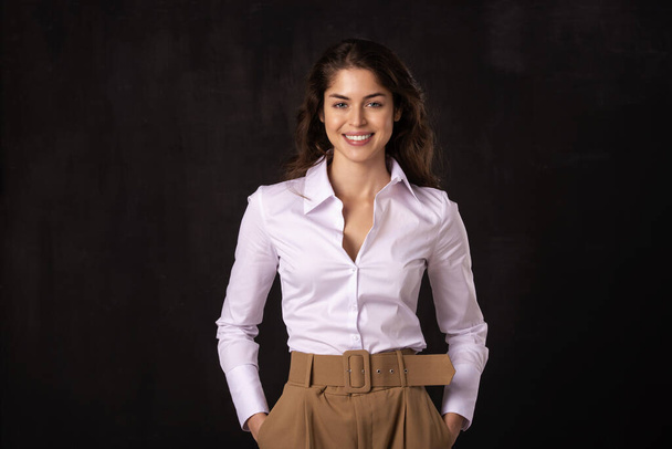 Portrait shot of attractive young woman wearing white shirt and laughing while standing at dark background.  - Foto, immagini